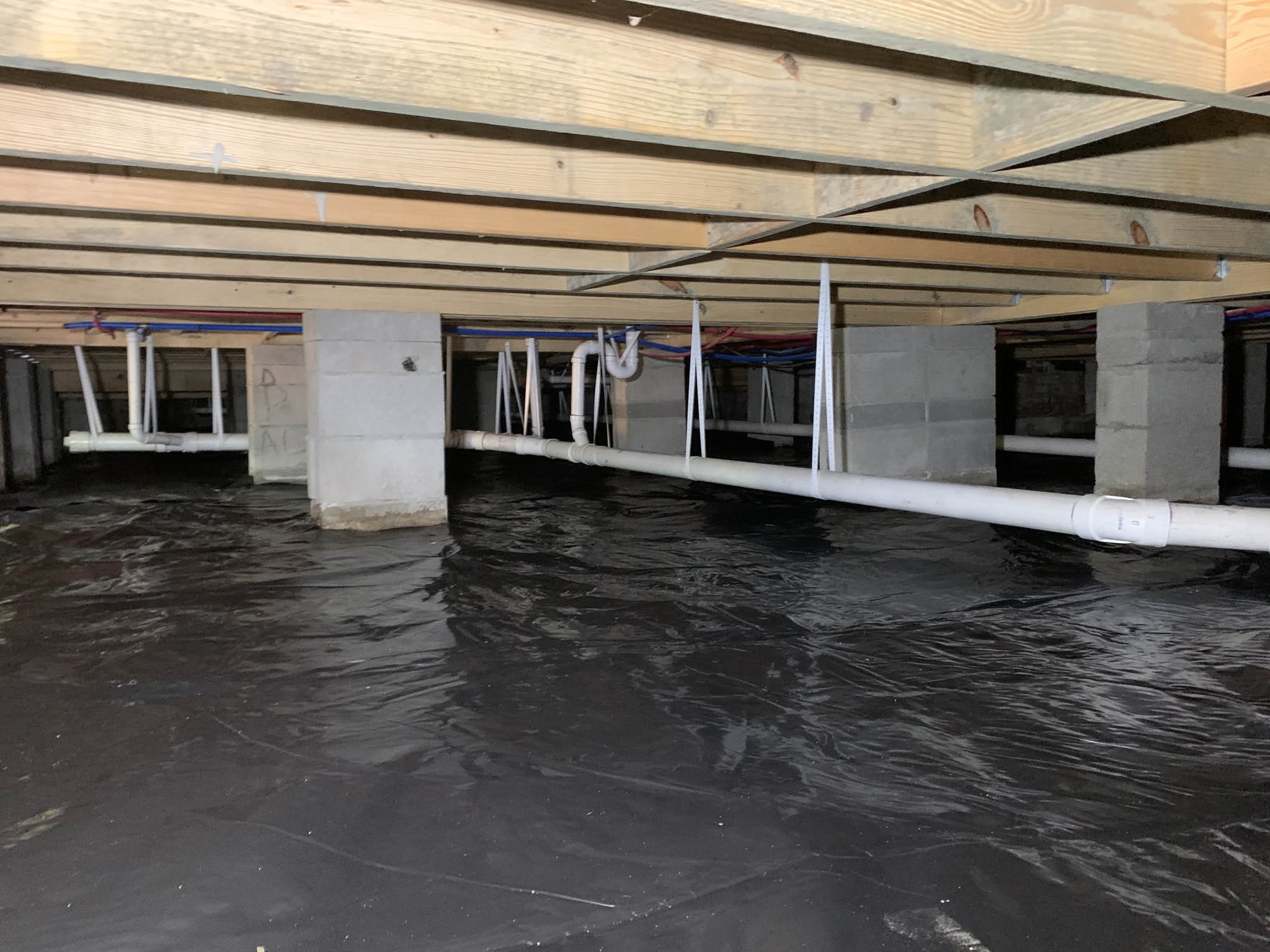 Crawl space with plastic vapor barrier on the ground, exposed pipes, concrete block supports, and wooden floor joists above.