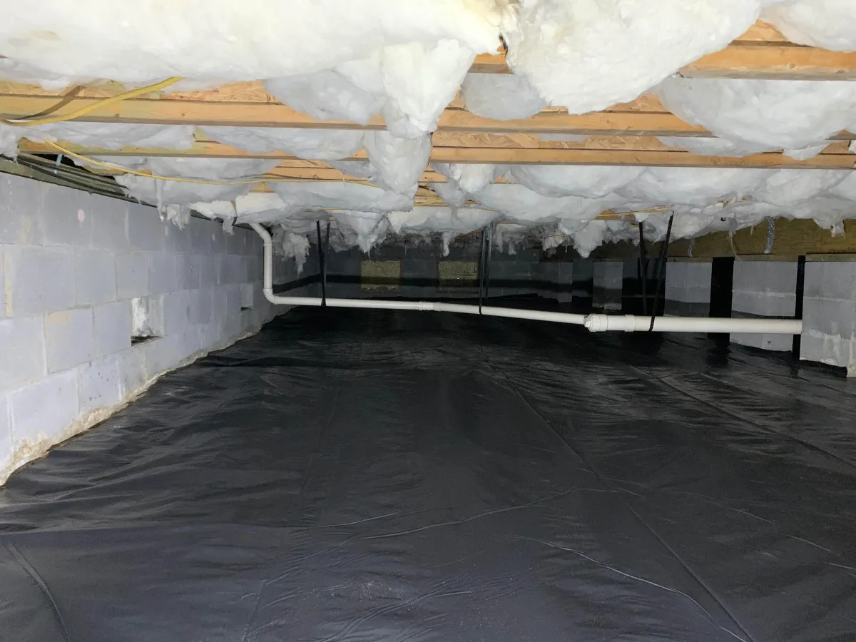 A crawl space with exposed insulation on the ceiling, plastic vapor barrier on the ground, concrete block walls, and visible white PVC pipes.
