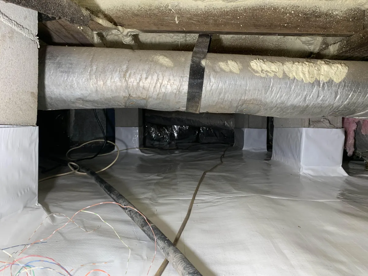 A crawl space with plastic vapor barrier on the floor and walls, exposed ductwork, cables, and concrete block supports.