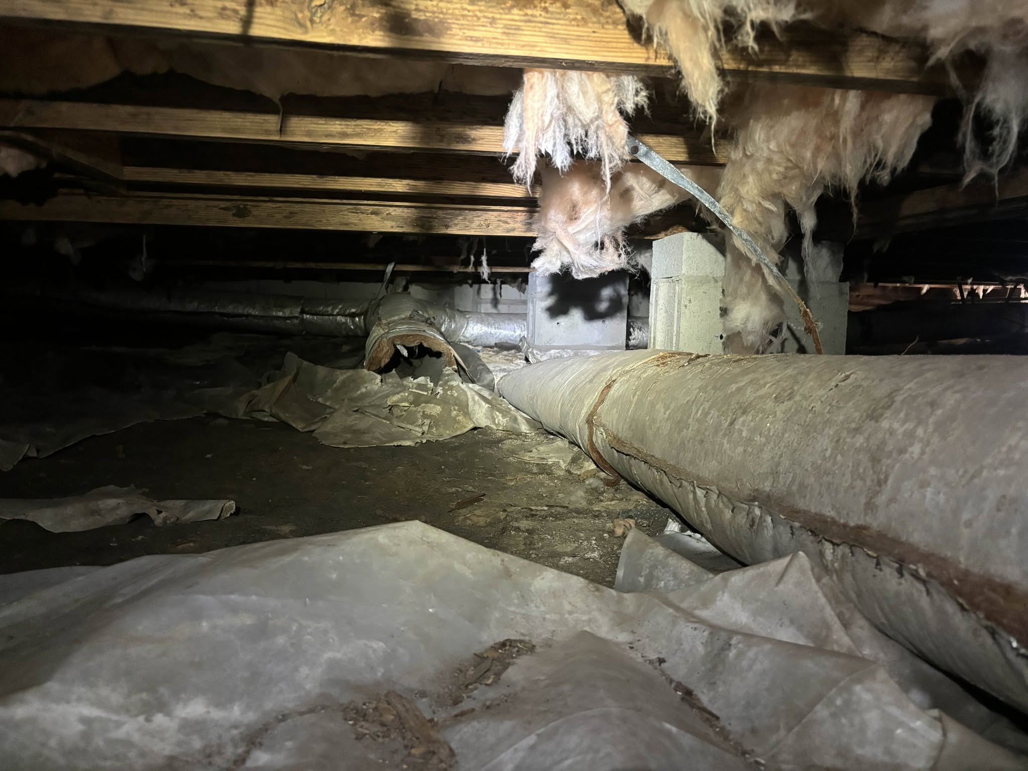A crawl space with exposed pipes, insulation hanging from the ceiling, and plastic sheeting covering the dirt floor.