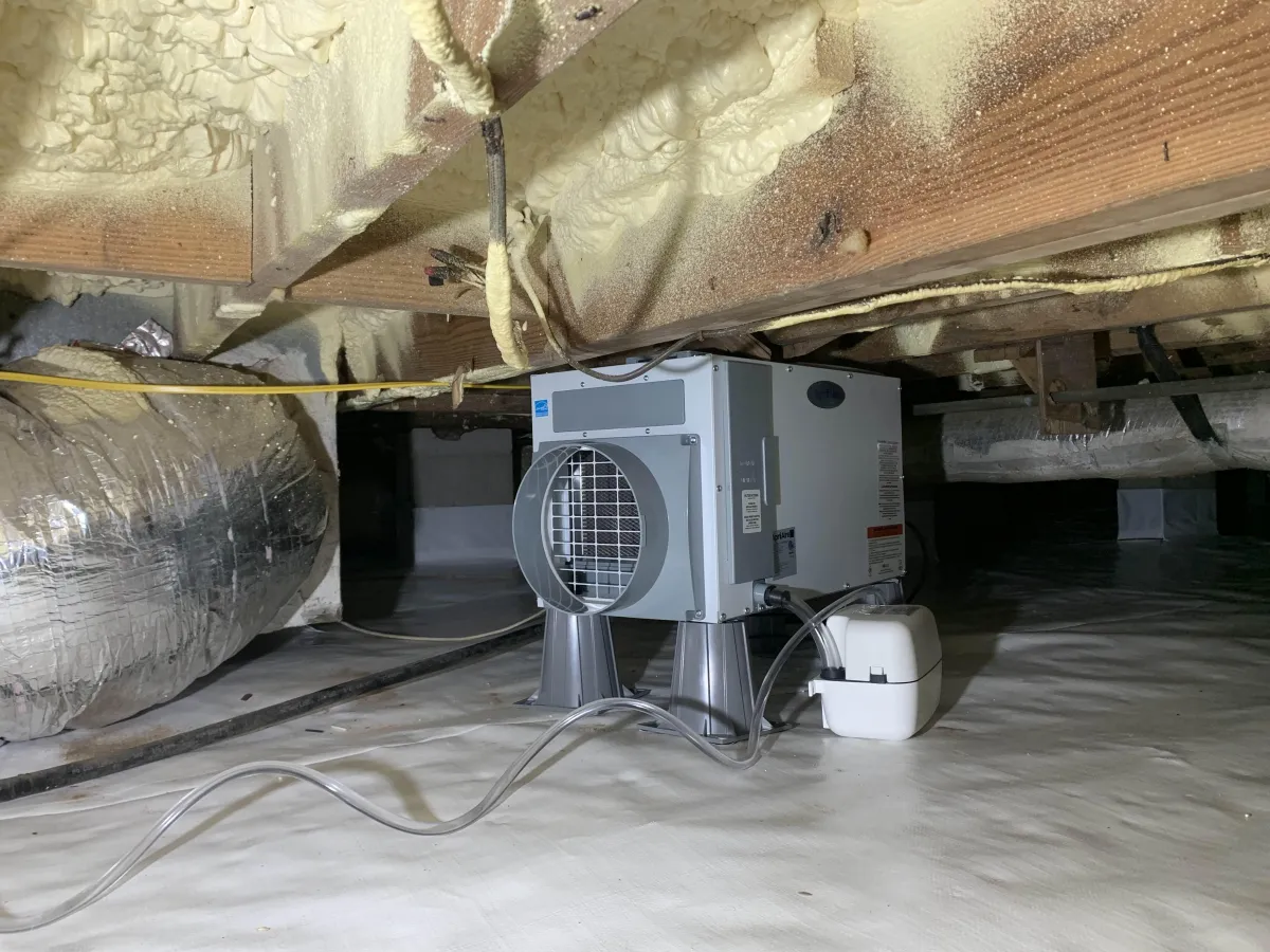 A dehumidifier unit is installed in a crawl space with foam insulation on the ceiling and a vapor barrier covering the floor.