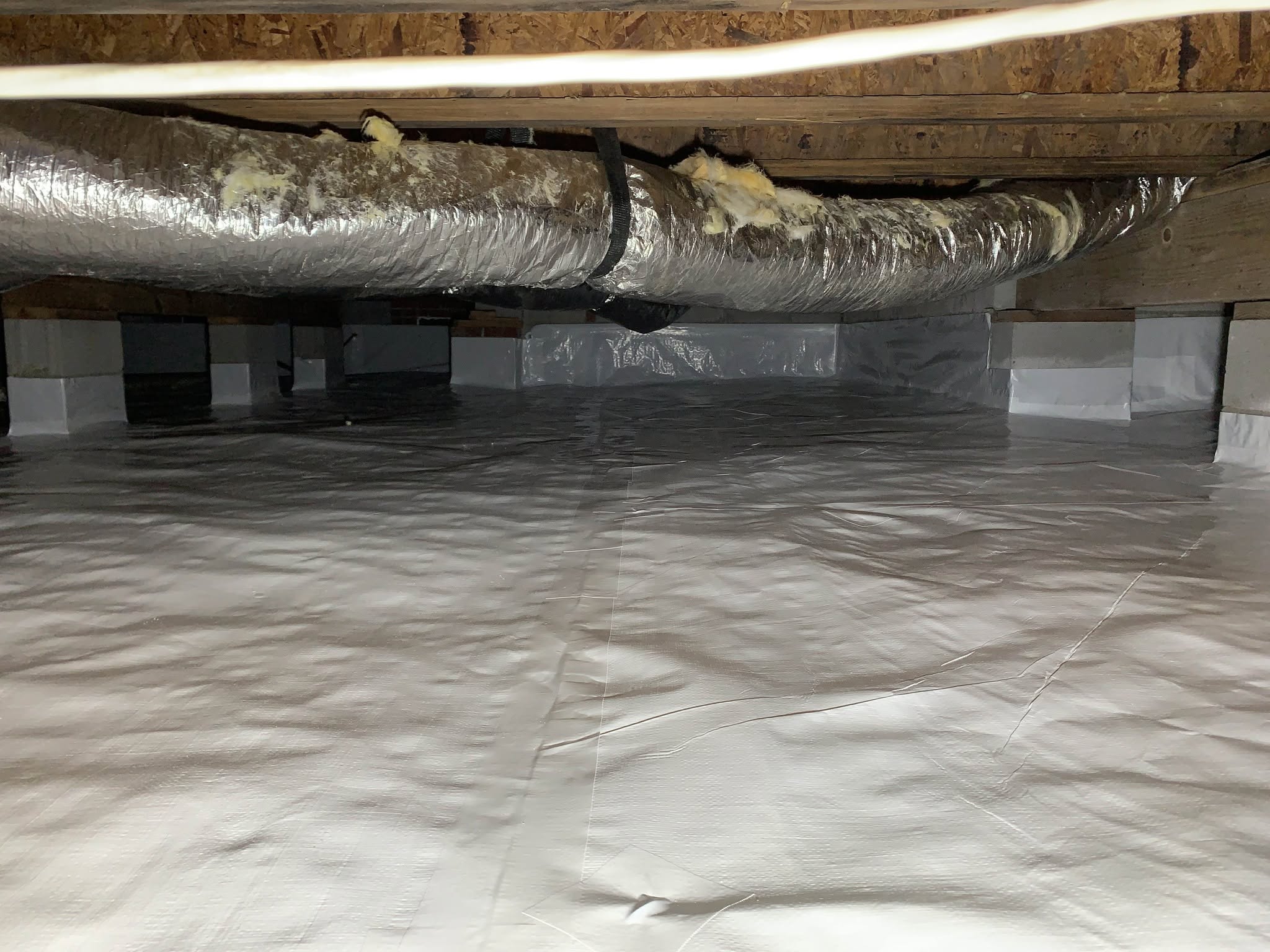 A crawl space with a clean, white vapor barrier covering the floor and walls, exposed ductwork above, and wooden beams supporting the ceiling.