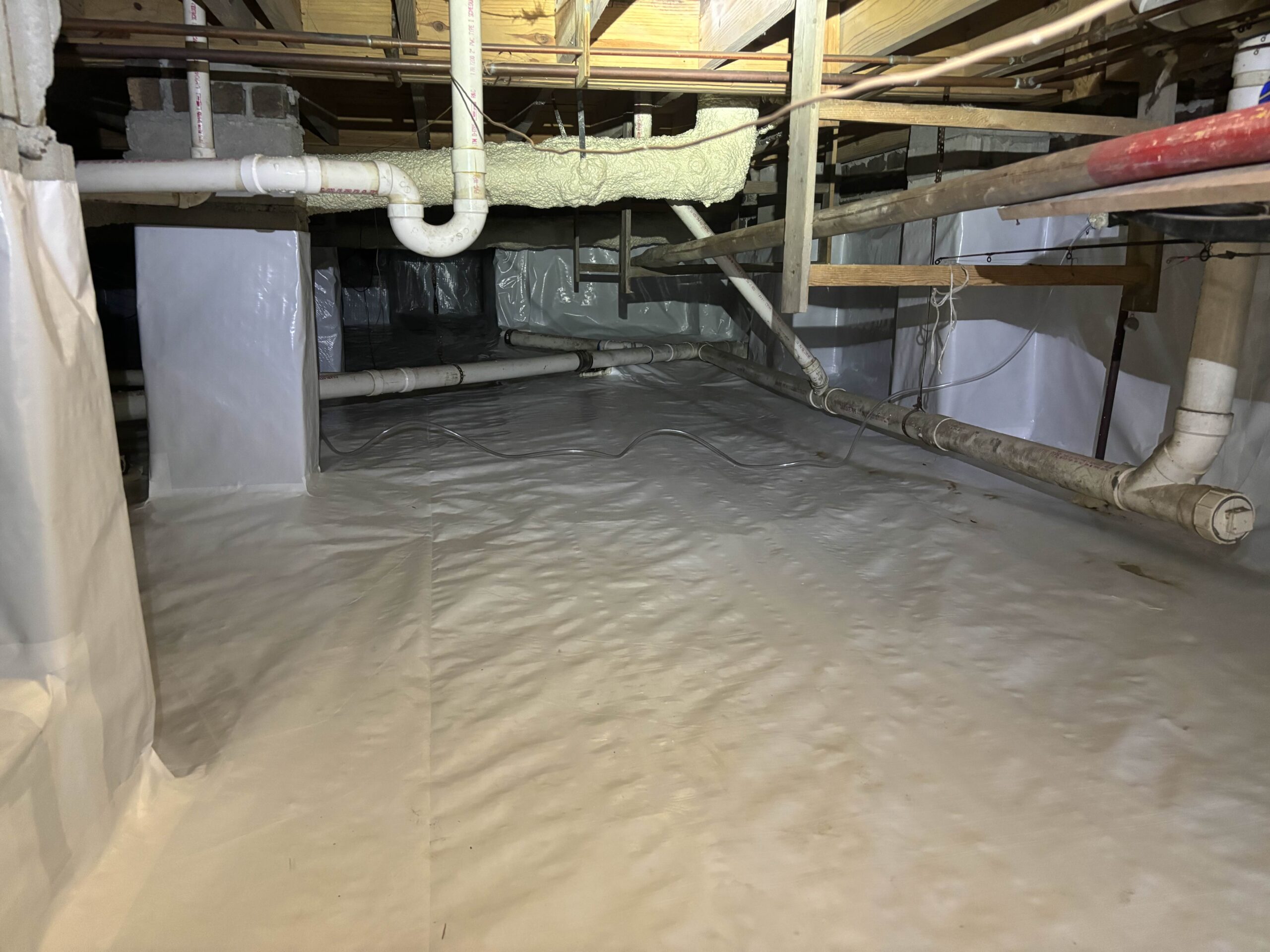 A crawl space with exposed pipes and wiring, lined with white plastic sheeting on the floor and walls for moisture control.