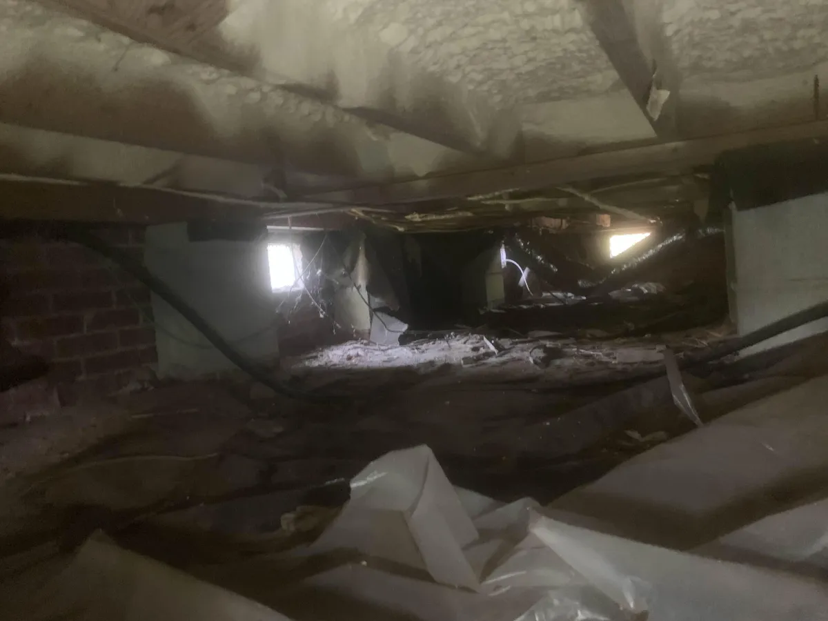 Dimly lit crawl space with exposed insulation, debris, and visible wiring, illuminated by small windows on one side.