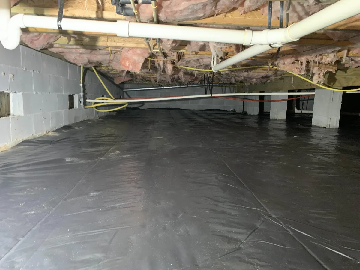 A crawl space with exposed insulation, pipes, and wiring above, and a black plastic vapor barrier covering the ground.