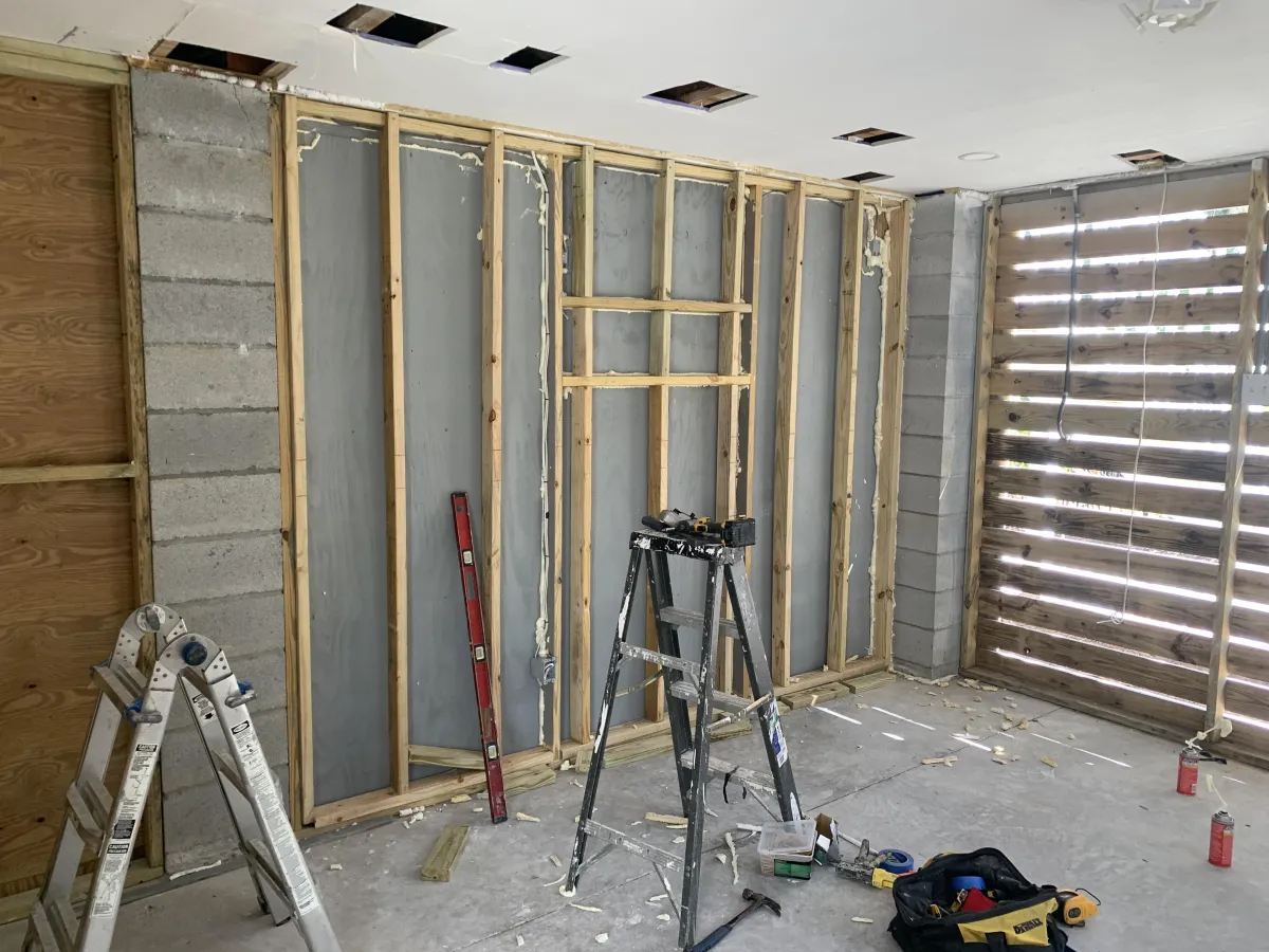 A room under construction with exposed wooden wall framing, tools, ladders, and construction materials scattered on the unfinished floor.