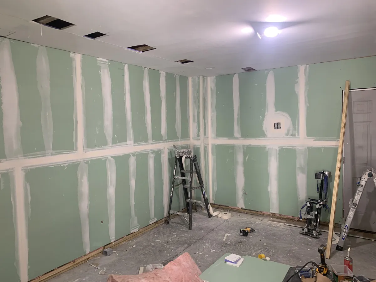 Unfinished room with green drywall panels, visible joint compound seams, scattered tools, a step ladder, and construction materials on the floor. Ceiling has open cutouts.