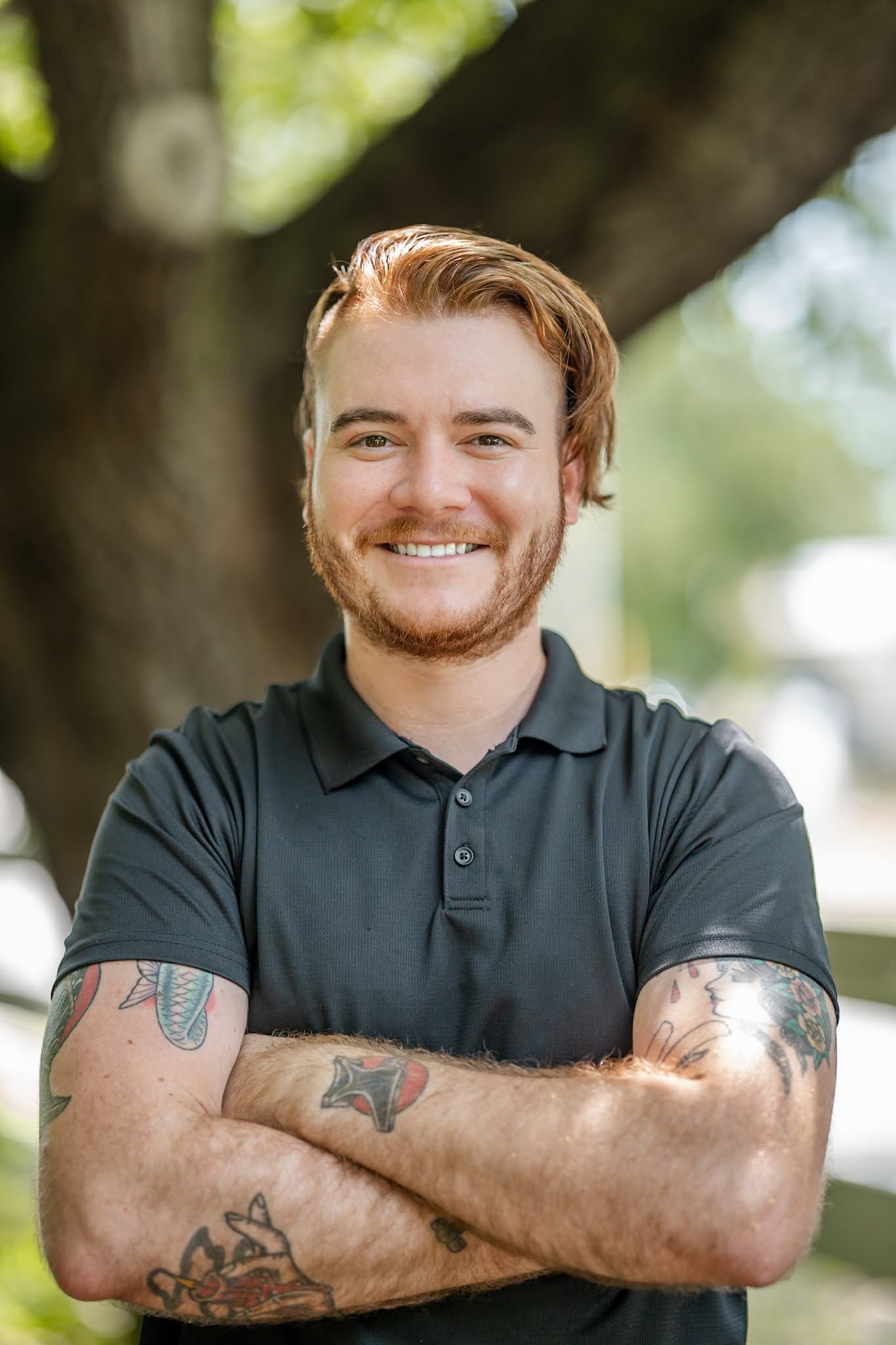 A person with light brown hair and a beard, wearing a dark polo shirt, stands outdoors with arms crossed, revealing several arm tattoos. A large tree is visible in the background.